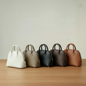 Puede incluir: Cinco bolsos pequeños en forma de cúpula con asas superiores y cierres de cremallera. Las bolsas se muestran en una fila, en tonos de blanco, beige, negro, marrón oscuro y marrón claro. Se colocan sobre una superficie de madera.