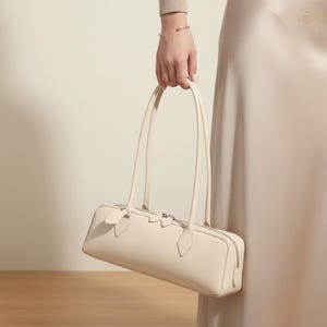 May include: A cream-colored rectangular handbag with long, thin handles. The bag features a zipper closure and decorative accents on the front. The bag is held by a person, showcasing its design and shape. The bag is made of a smooth material.