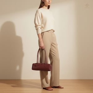 May include: A burgundy leather handbag with a rectangular shape and long handles. The bag is held by a person wearing a cream-colored sweater and tan pants. The bag has a decorative element on the front.