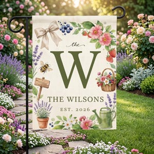 May include: A rectangular garden flag featuring a large olive green letter W, the text the, THE WILSONS, and EST. 2026. The design includes a beige bow, blueberries, pink flowers, strawberries, a bee, a wooden sign, a basket, and a green watering can.