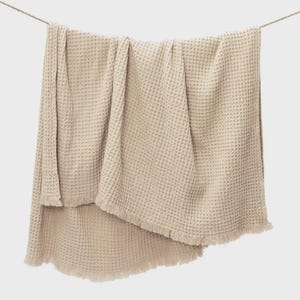 May include: A beige waffle-weave blanket with a fringed edge is draped over a clothesline. The blanket is made of a soft, textured material and hangs in soft folds. The neutral color and simple design make it suitable for various home decor styles.