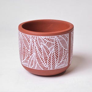 May include: A terracotta-colored ceramic plant pot with a white leaf pattern. The cylindrical pot has a rounded base and a wide opening. The white leaf design covers the lower portion of the pot, creating a contrast against the reddish-brown background.