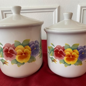 Puede incluir: Dos botes de cerámica blanca con tapas. Cada bote está decorado con un diseño floral con pensamientos rojos, amarillos y morados con hojas verdes. Los botes están sobre una superficie roja.