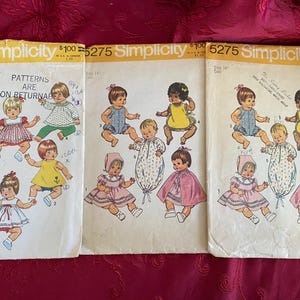 May include: Three vintage Simplicity sewing pattern envelopes for dolls, numbered 5947 and 5275. The white paper envelopes feature illustrations of dolls wearing various outfits, including dresses, rompers, and capes, with printed text and pricing.