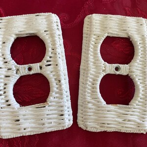 May include: Two white wicker-style electrical outlet covers. Each cover has two circular openings for the outlets and a rectangular shape. The covers are made of woven material and have a textured appearance. The background is a red patterned fabric.