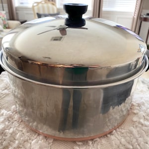 May include: Stainless steel cooking pot with a lid and black handles. The pot has a hammered texture and a copper-colored base. The lid has a black knob. The pot is sitting on a white, textured surface.