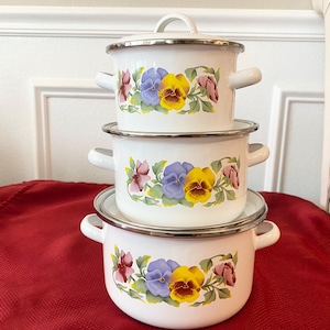 May include: Three white enamel pots with lids and handles are stacked. Each pot is decorated with a floral design featuring yellow, purple, and pink pansies. The pots are arranged on a red tablecloth.