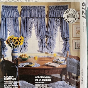 Op de afbeelding: Een McCalls Home Decorating naaipatroon envelop 6727 met blauw-witte gestreepte gordijnen met ruches, bijpassende stoelkussens en een gedekte tafel. De tekst bevat KITCHEN ESSENTIALS en LES ESSENTIELS DE LA CUISINE.