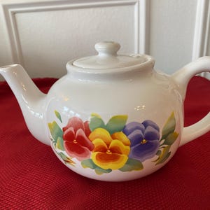 Op de afbeelding: Witte keramische theepot met een deksel en handvat. De theepot is versierd met handgeschilderde bloemen in rood, geel en paars, met groene bladeren. De theepot staat op een rode ondergrond.