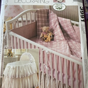Op de afbeelding: Een McCall's Home Decorating patroon voor een babybedset. Het patroon toont een wieg met een roze en witte geruite deken, een bijpassende bumper en een teddybeer en konijn. Het patroon bevat ook een wieg.