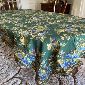 May include: A green floral tablecloth featuring a pattern of blue, yellow, and red flowers. The tablecloth covers an oval table. The edges are trimmed with a burgundy border.