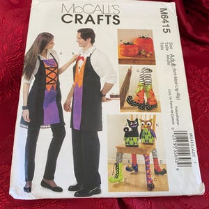 May include: A McCall's Crafts M6415 sewing pattern envelope for adult costumes. The front displays illustrations of aprons with orange and purple accents, decorative fabric pumpkins, striped witch legs, and plush cat and owl figures.