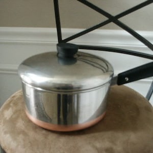 May include: A stainless steel saucepan with a copper-colored base, a matching stainless steel lid with a black knob, and a black handle. The pot features a polished metallic finish and a circular design, providing a functional kitchen tool.