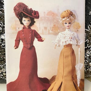May include: A vintage Butterick sewing pattern featuring two dolls dressed in period clothing. One doll wears a burgundy coat dress and feathered hat, the other a mustard-colored skirt and lace top. The pattern is labeled "63/4" and "ONE SIZE".