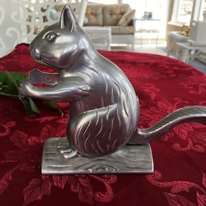 Könnte beinhalten: Ein silberfarbener Eichhörnchen-Nussknacker. Das Eichhörnchen sitzt auf einem Holzsockel und hält eine Nuss. Der Hintergrund ist eine rote Tischdecke mit floralem Muster.