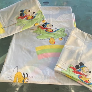 Puede incluir: Juego de sábanas blancas con Mickey Mouse en un avión verde con capa roja y Pluto. El juego incluye una sábana encimera, una sábana bajera y una funda de almohada con un diseño de arcoíris colorido.