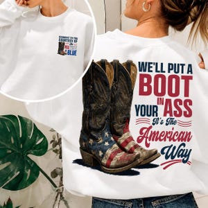 Puede incluir: Una sudadera blanca de manga larga con un gráfico de dos botas vaqueras con patrones de la bandera estadounidense. El texto trasero dice: WE'LL PUT A BOOT IN YOUR ASS It's The American Way. El pecho tiene un gráfico más pequeño a juego.