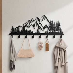 May include: Black metal wall-mounted coat rack with a mountain and forest silhouette design. The rack has multiple hooks and is holding a light gray towel, a beige handbag, a teddy bear keychain, a key ring, and a beige coat.