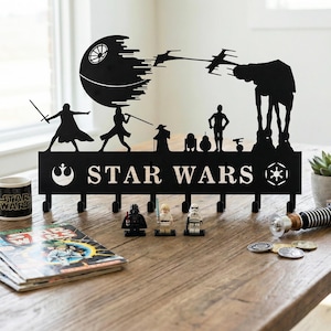 May include: Black metal Star Wars key holder featuring silhouettes of characters and the Death Star. Includes hooks for keys, a Star Wars mug, comic books, and figurines on a wooden table.