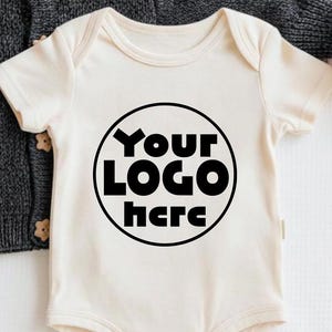 Może przedstawiać: Kremowe body niemowlęce z napisem "Your LOGO here" w czarnym okręgu. Za body znajduje się ciemnoszary dzianinowy kardigan z drewnianymi guzikami. Po prawej stronie znajduje się pomarańczowa dekoracja w kształcie dyni.