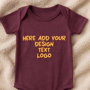 May include: A maroon baby onesie with short sleeves and snap closures. The text "HERE ADD YOUR DESIGN TEXT LOGO" is printed in yellow across the chest. The onesie is displayed on a cream-colored, textured surface.
