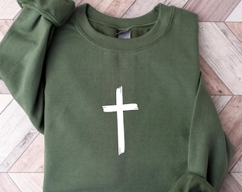 May include: An olive green sweatshirt with a white, hand-painted cross design. The long-sleeved top has a crew neck and ribbed cuffs. The sweatshirt is made of a soft material and is suitable for casual wear.