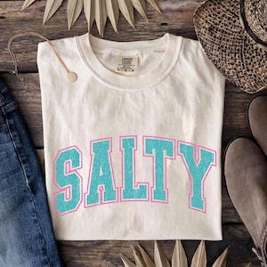 May include: Cream-colored t-shirt with the word "SALTY" in large, turquoise block letters outlined in pink. The shirt is made of a soft material and has a classic crew neck. The shirt is displayed on a wooden surface.