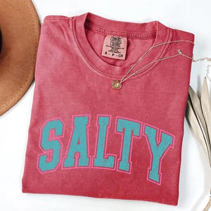 May include: A red Comfort Colors t-shirt with the word "SALTY" in teal block letters outlined in pink. A gold necklace with a pendant rests on the shirt. The shirt is folded and displayed on a white surface.