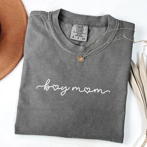 May include: A gray Comfort Colors t-shirt with the words "boy mom" in white script. A silver necklace with a pendant is draped across the shirt. A brown hat and dried palm leaves are in the background.