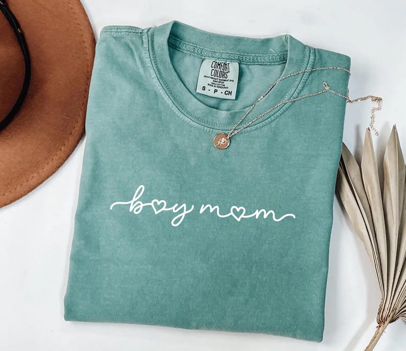 May include: A seafoam green t-shirt with the words "boy mom" in white script. A silver necklace with a small pendant rests on the shirt. The shirt is folded, and a brown hat and dried plant are in the background.