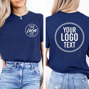 Pu&ograve; includere: Una t-shirt a maniche corte blu navy con una grafica circolare bianca sul petto sinistro e una versione pi&ugrave; grande della stessa grafica sul retro. La grafica contiene il testo YOUR LOGO TEXT in un carattere sans-serif in grassetto all'interno di un doppio cerchio.