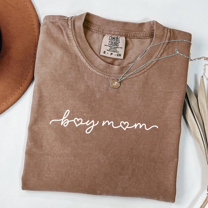 May include: A brown Comfort Colors t-shirt with the words "boy mom" in white script. A silver necklace with a small pendant is draped across the shirt. The shirt is size small.