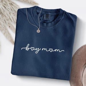 May include: Navy blue t-shirt with the words "boy mom" in white script, with a heart replacing the "o". A silver necklace with a pendant rests on the shirt. The shirt is a size L-G-G.
