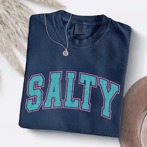 May include: A navy blue t-shirt with the word "SALTY" in large, distressed teal letters outlined in pink. A silver necklace with a round pendant rests on the shirt. The shirt is folded neatly.