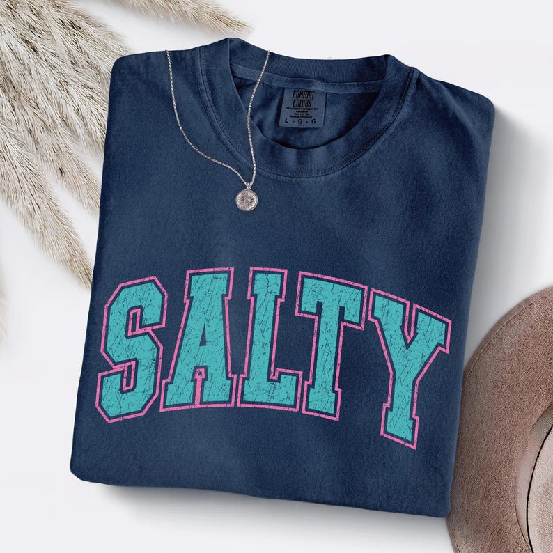 May include: A navy blue t-shirt with the word "SALTY" in large, distressed teal letters outlined in pink. A silver necklace with a round pendant rests on the shirt. The shirt is folded neatly.