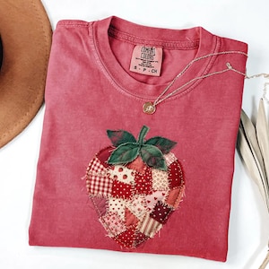 May include: A red Comfort Colors t-shirt with a patchwork strawberry design. The strawberry is made of various red and white patterned fabrics with green leaves. A gold necklace with a small pendant rests on the shirt.