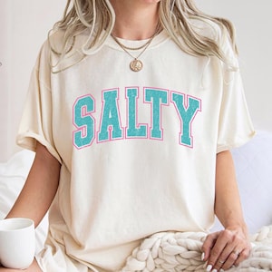 May include: Cream-colored t-shirt with the word "SALTY" in turquoise block letters outlined in pink. The shirt has a relaxed fit and is being worn.