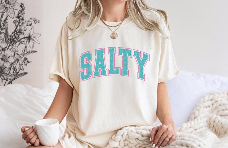 May include: Cream-colored t-shirt with the word "SALTY" in turquoise block letters outlined in pink. The shirt has a relaxed fit and is being worn.