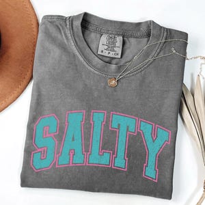 May include: A gray Comfort Colors t-shirt with the word "SALTY" in turquoise block letters outlined in pink. A silver necklace with a small pendant rests on the shirt. A brown hat is visible in the upper left corner.
