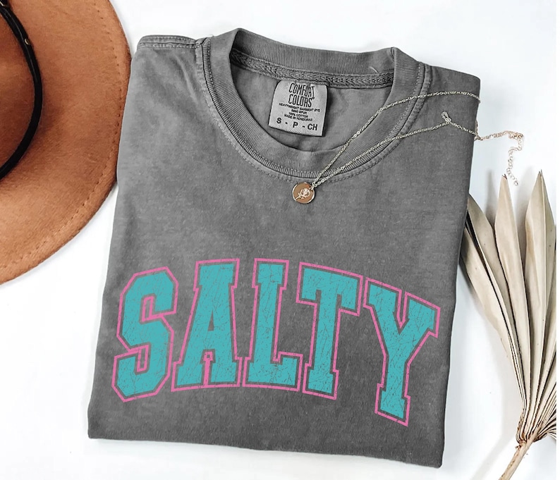 May include: A gray Comfort Colors t-shirt with the word "SALTY" in turquoise block letters outlined in pink. A silver necklace with a small pendant rests on the shirt. A brown hat is visible in the upper left corner.