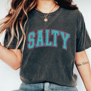 May include: Dark gray t-shirt with the word "SALTY" in large, turquoise block letters outlined in pink. The shirt has a vintage, worn look, and is a casual, comfortable style.