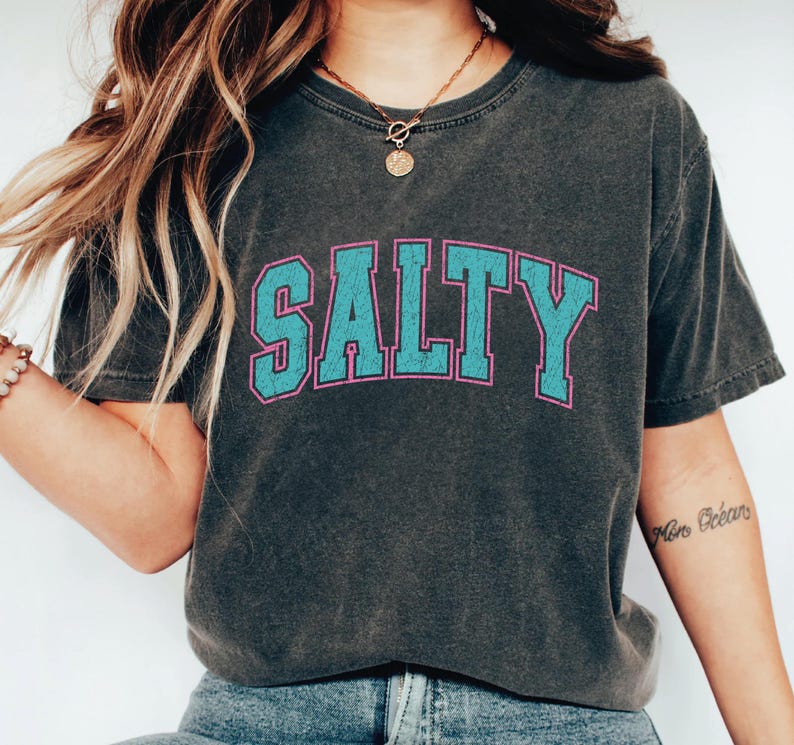 May include: Dark gray t-shirt with the word "SALTY" in large, turquoise block letters outlined in pink. The shirt has a vintage, worn look, and is a casual, comfortable style.