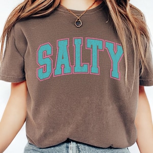 May include: A brown t-shirt with the word "SALTY" printed across the front in turquoise letters with a pink outline. The shirt is a crew neck and appears to be made of a soft material.