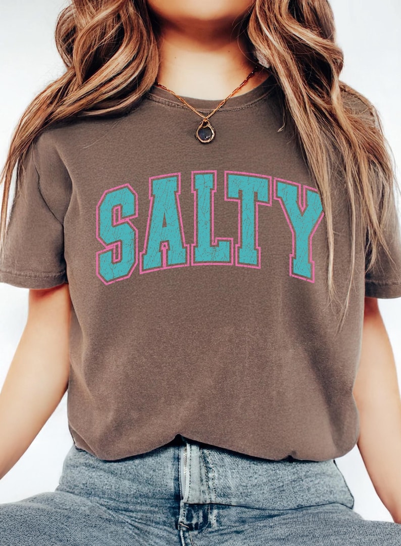 May include: A brown t-shirt with the word "SALTY" printed across the front in turquoise letters with a pink outline. The shirt is a crew neck and appears to be made of a soft material.