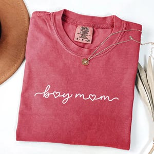 May include: A red t-shirt with the words "boy mom" in white script. A gold necklace with a pendant is draped over the shirt. The shirt is folded, and a brown hat is visible in the background.
