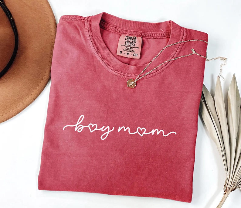 May include: A red t-shirt with the words "boy mom" in white script. A gold necklace with a pendant is draped over the shirt. The shirt is folded, and a brown hat is visible in the background.
