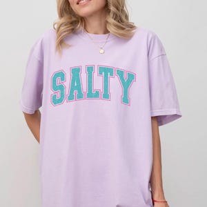 May include: A light purple t-shirt with the word "SALTY" in teal block letters with a white outline. The shirt is a relaxed fit and has short sleeves. The overall style is casual and comfortable.