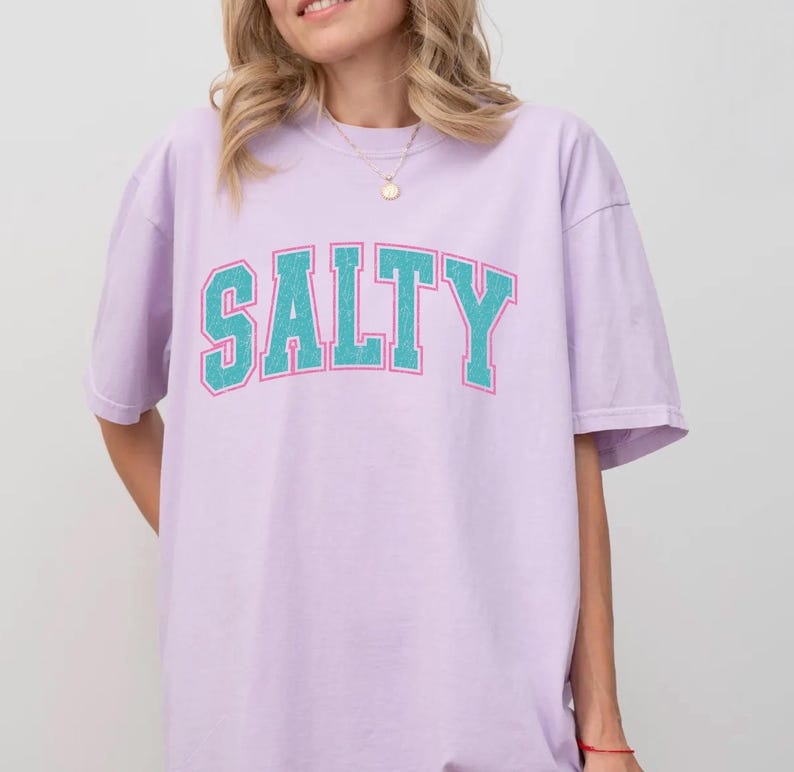 May include: A light purple t-shirt with the word "SALTY" in teal block letters with a white outline. The shirt is a relaxed fit and has short sleeves. The overall style is casual and comfortable.