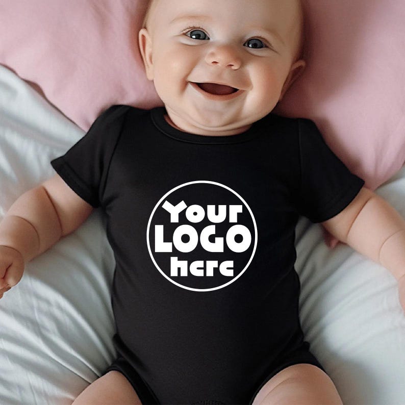Może przedstawiać: Czarny body niemowlęce z kr&oacute;tkim rękawem. Body ma białe okrągłe logo z napisem "Your LOGO here". Dziecko uśmiecha się i leży na r&oacute;żowej poduszce.