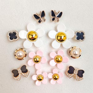 Op de afbeelding: Een verzameling decoratieve versieringen, waaronder vlinder-, bloem-, strik- en klaverontwerpen. De versieringen hebben gouden, zwarte, witte, roze en parelaccenten. Deze decoratieve stukken zijn geschikt voor diverse knutselprojecten.